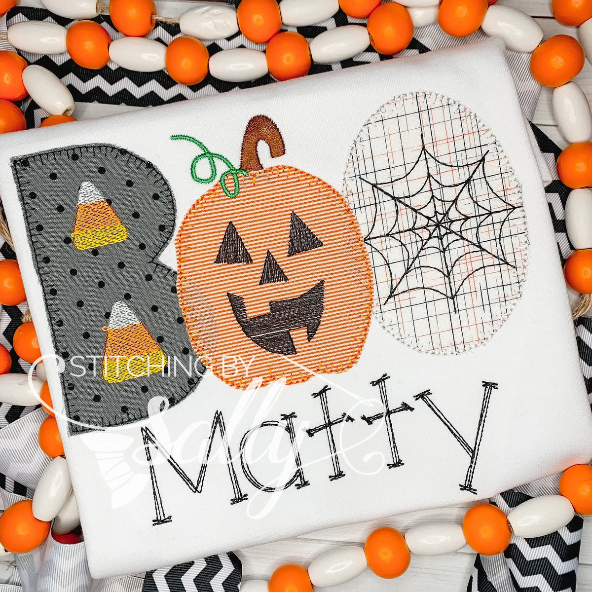 A white shirt with a Halloween-themed applique design including a pumpkin, candy corn, and spider web.
