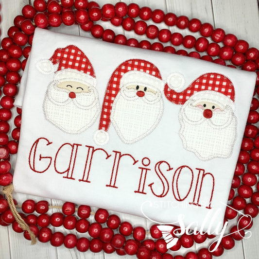 A white shirt with a santa trio appliqué in red and white fabric. A name is embroidered in read thread underneath.
