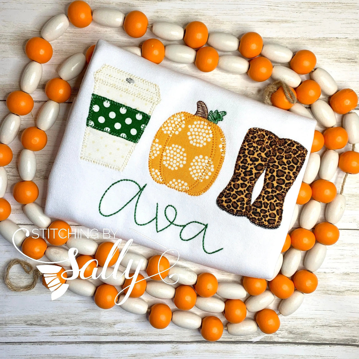 A white shirt with applique decorations featuring a latte cup, pumpkin, and leopard print boots, displayed on a surface with orange beads.