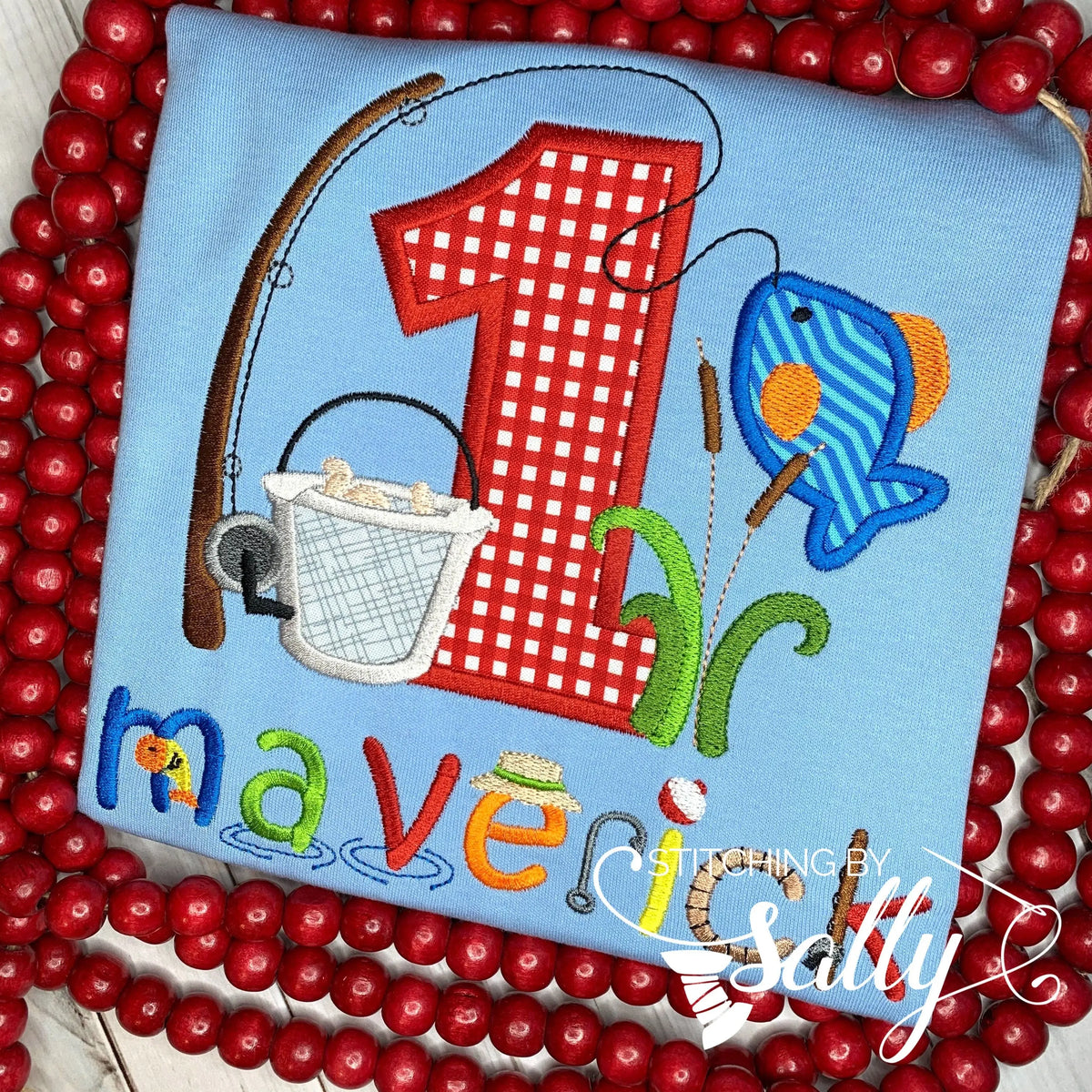 A blue shirt featuring a cute fishing lure and fish applique design in bright colors. That design is centered around a large 1 appliquéd in red gingham fabric. Embroidered underneath is a name in a fun fishing font.