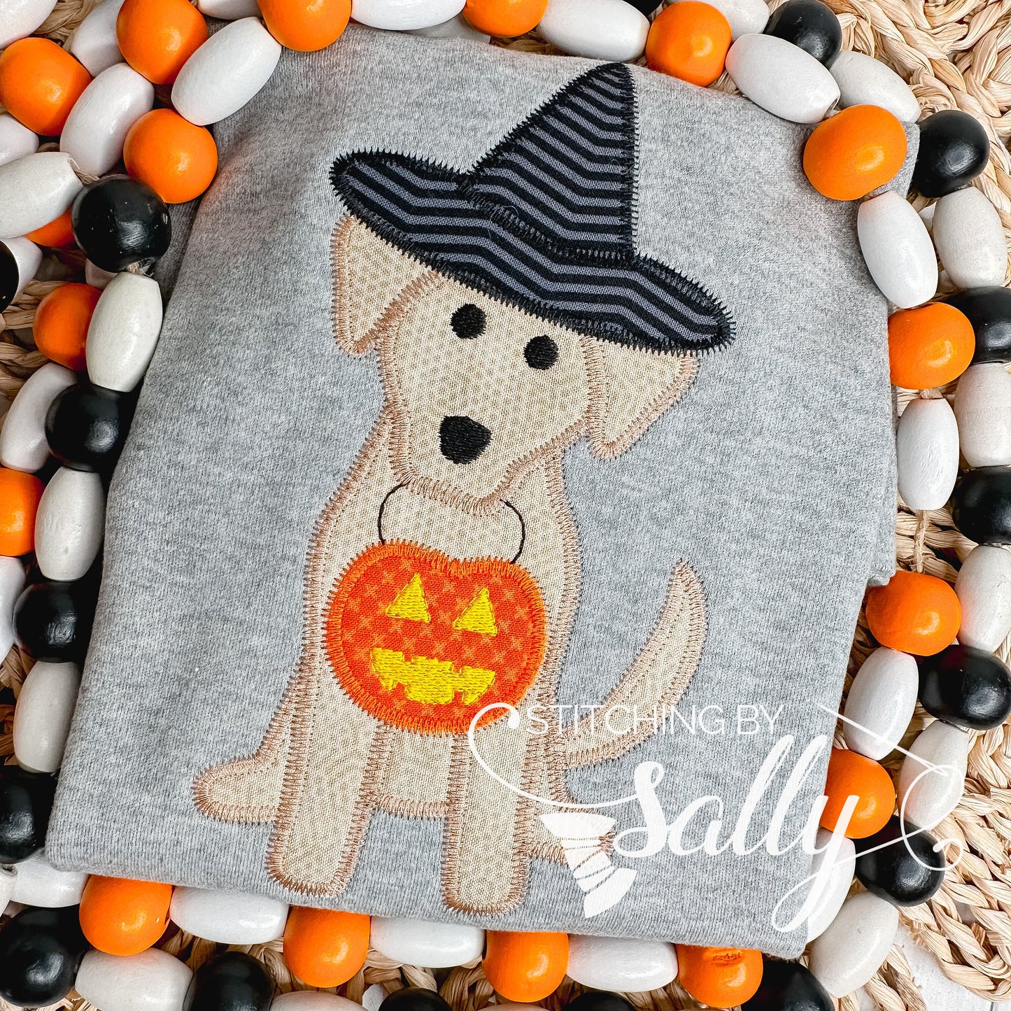 A gray shirt with an appliqué of a puppy wearing a witch's hat and holding a pumpkin. The shirt is displayed on a surface with decorative orange, black, and white beads.