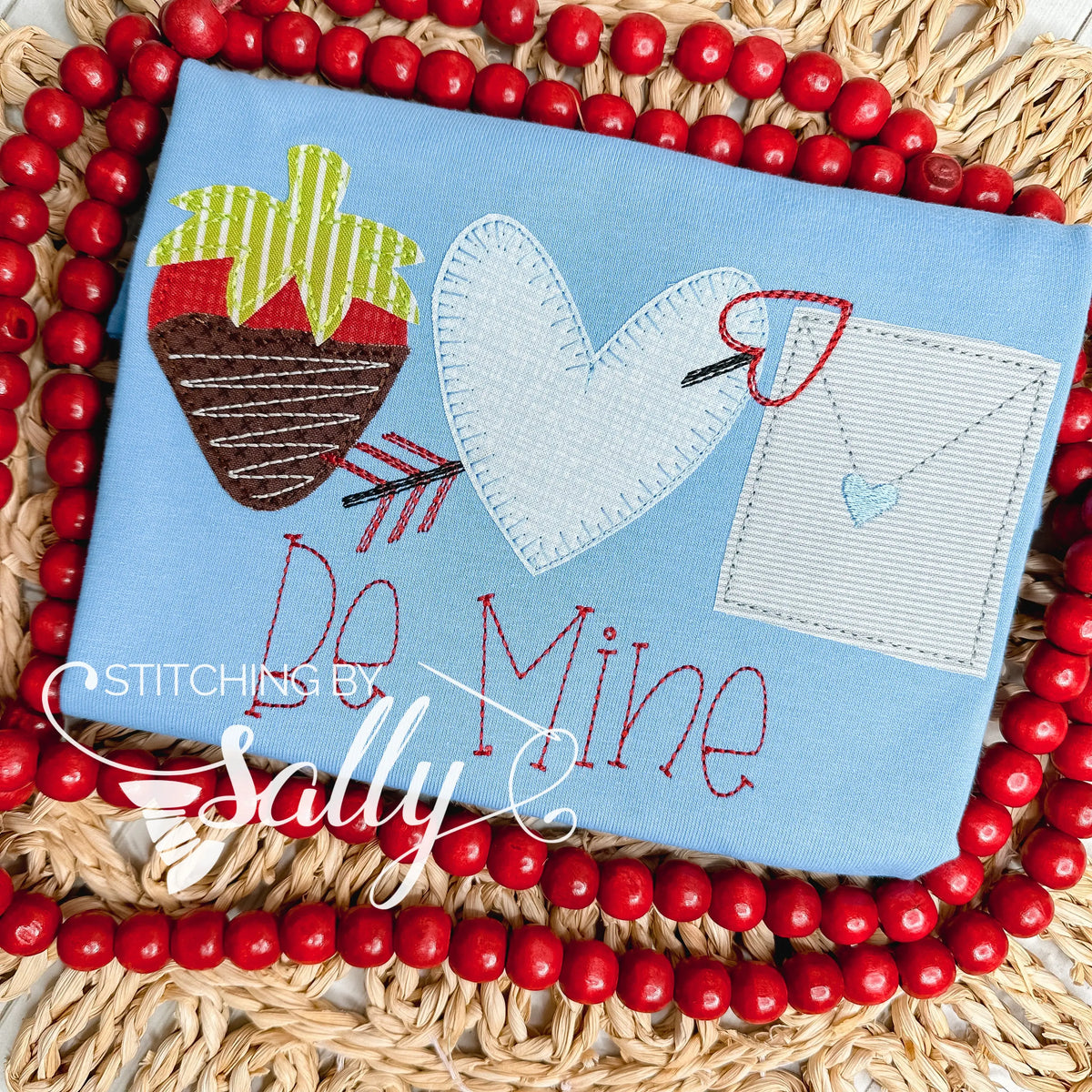 A strawberry, heart with arrow, and a letter are appliqué on a blue shirt. Be mine is embroidered underneath.
