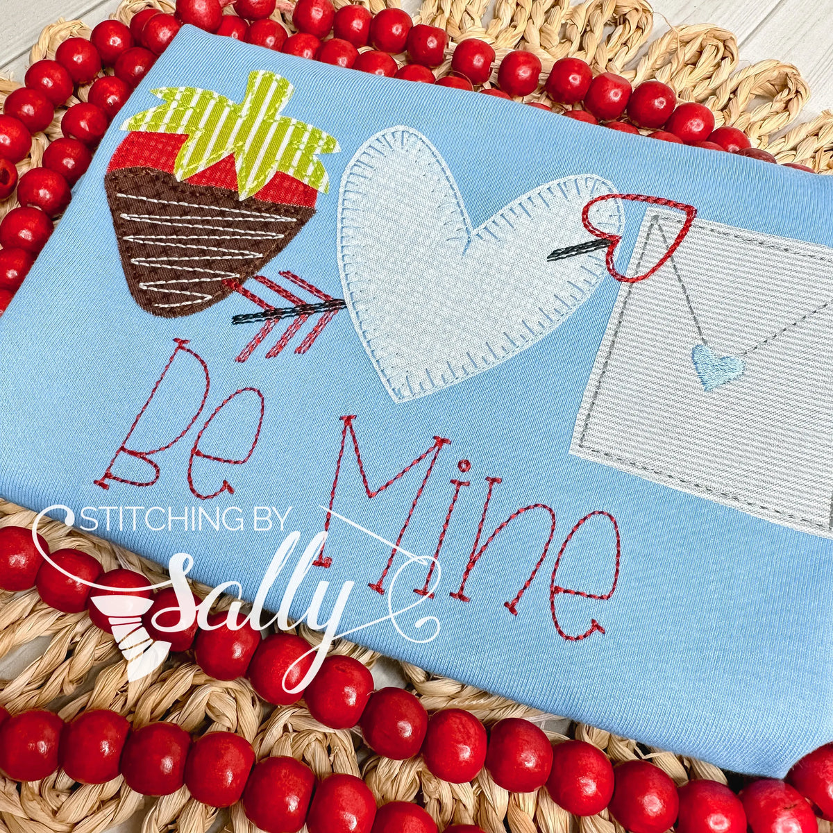 A blue shirt with a strawberry, heart with arrow, and letter appliqué designs. Be mine is embroidered underneath.
