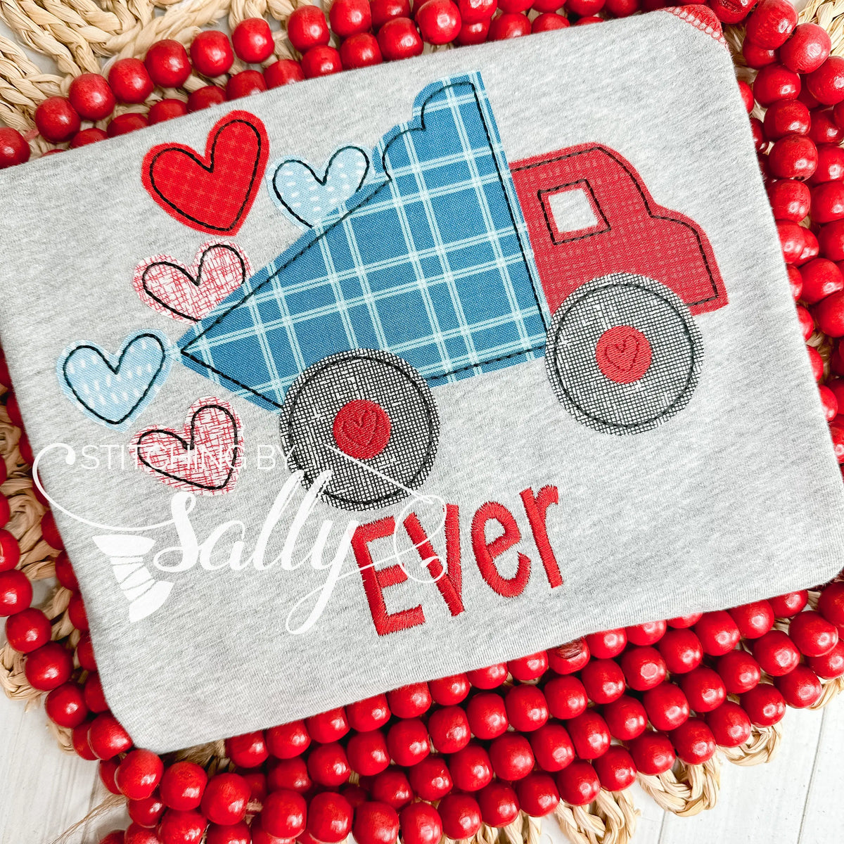 A grey shirt with a red and blue dump truck appliqué design. Truck is dumping red heart appliqués.