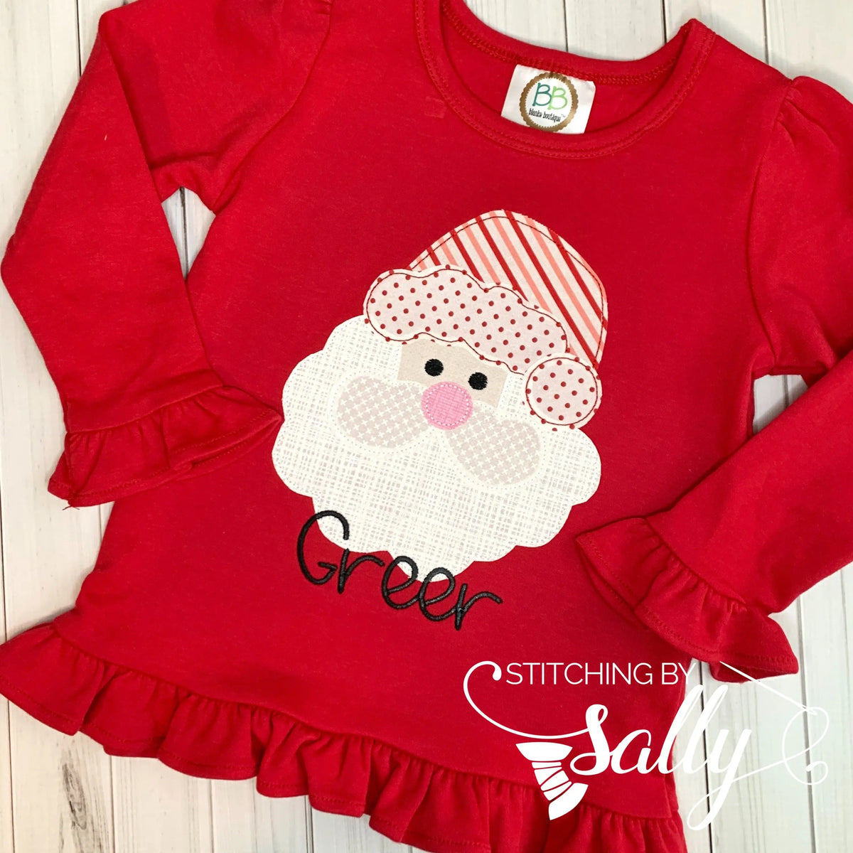 A long sleeve red shirt with ruffles with a santa face applique in red and white fabric. Personalized with name in black thread.