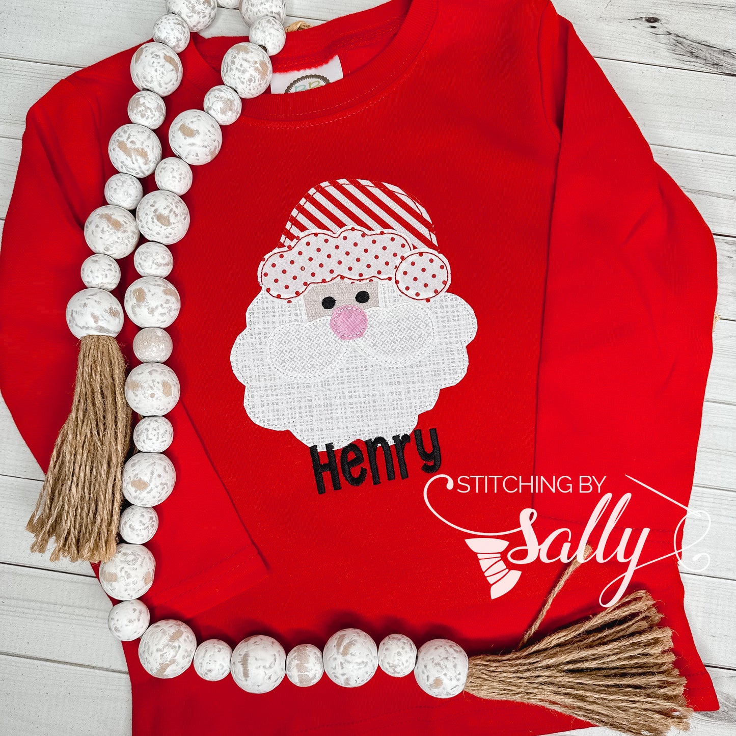 Red shirt with Santa Claus design and 'Henry' text, decorated with beads and tassels on a wooden surface.