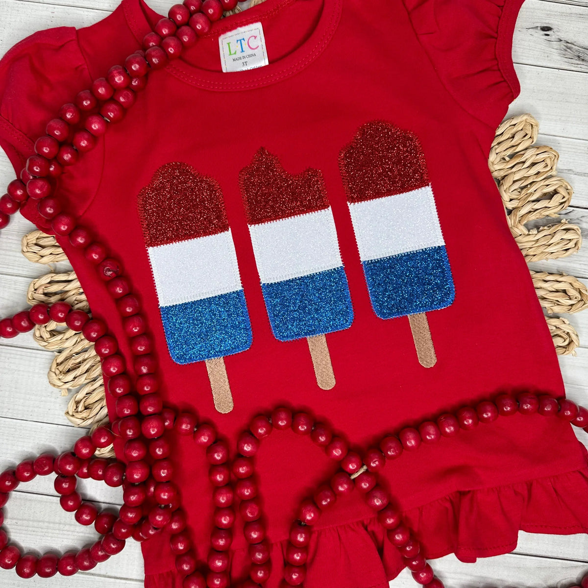 This red ruffle shirt features an applique design of the firecracker popsicle, in red, white and blue glitter htv.
