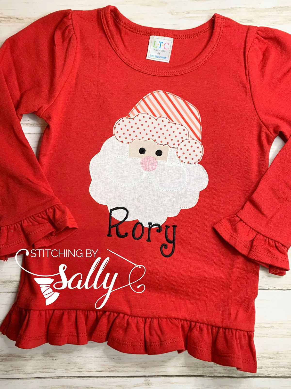 A long sleeve red shirt with ruffles with a santa face applique in red/pink and white fabric. Personalized with name in black thread.