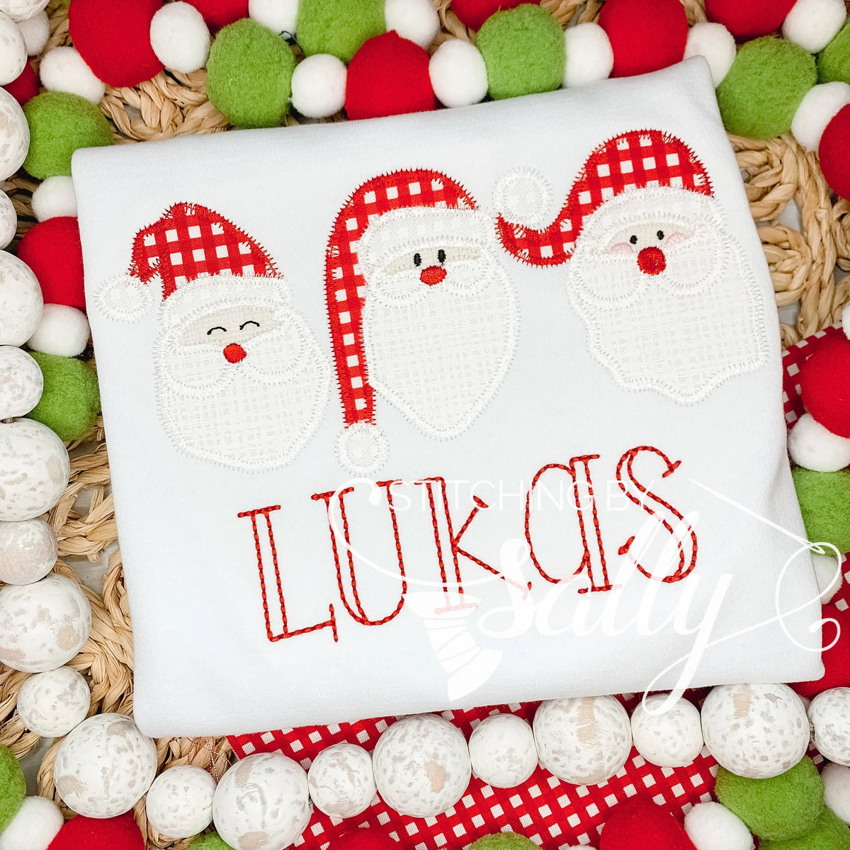 A white shirt with a santa trio appliqué in red and white fabric. A name is embroidered in read thread underneath.