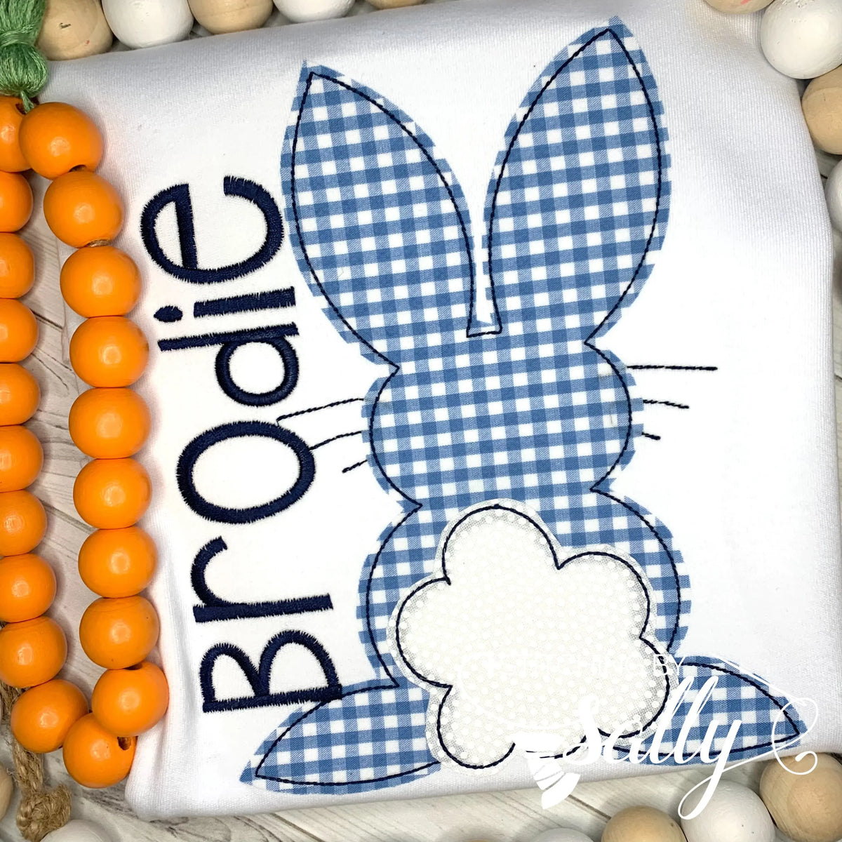 A white shirt featuring a blue gingham appliqué design of the Easter Bunny. A name is embroidered on the side