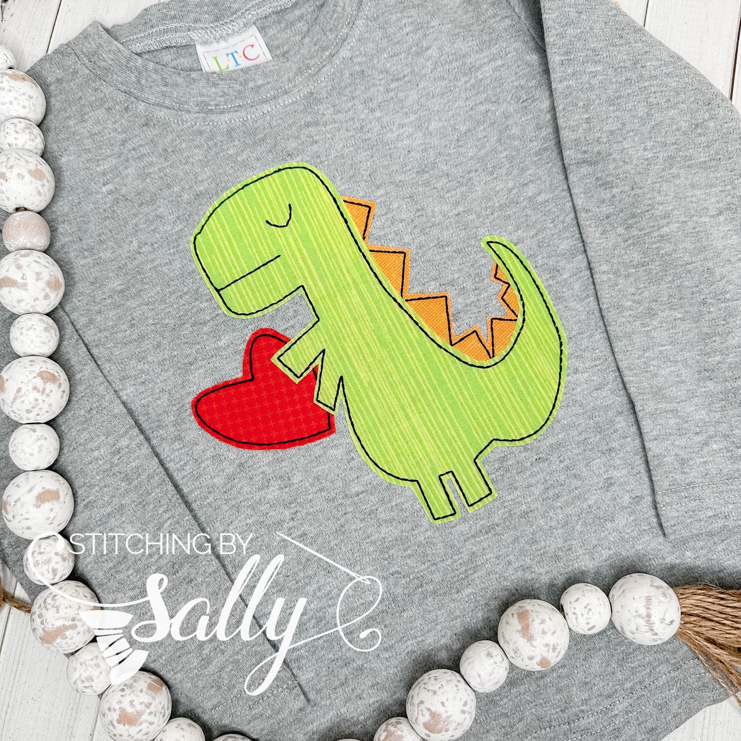 Boys grey long sleeve shirt with a green and orange dinosaur appliqué. Dinosaur is holding a red heart. 