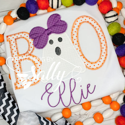 A white t-shirt with the word 'BOO' written in orange and black with a purple bow on the 'O'. The t-shirt is personalized with the name 'Ellie' in a purple script.