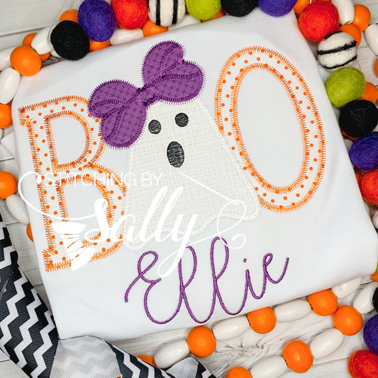 A white t-shirt with the word 'BOO' written in orange and black with a purple bow on the 'O'. The t-shirt is personalized with the name 'Ellie' in a purple script.