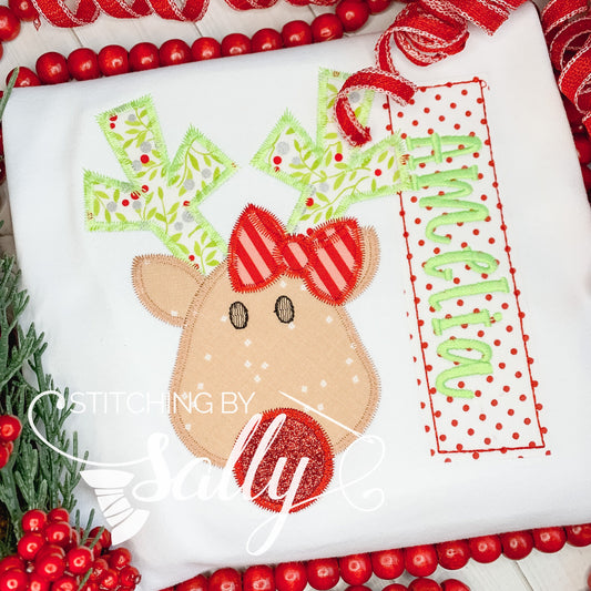 A white shirt with a reindeer applique design and a name embroidered.