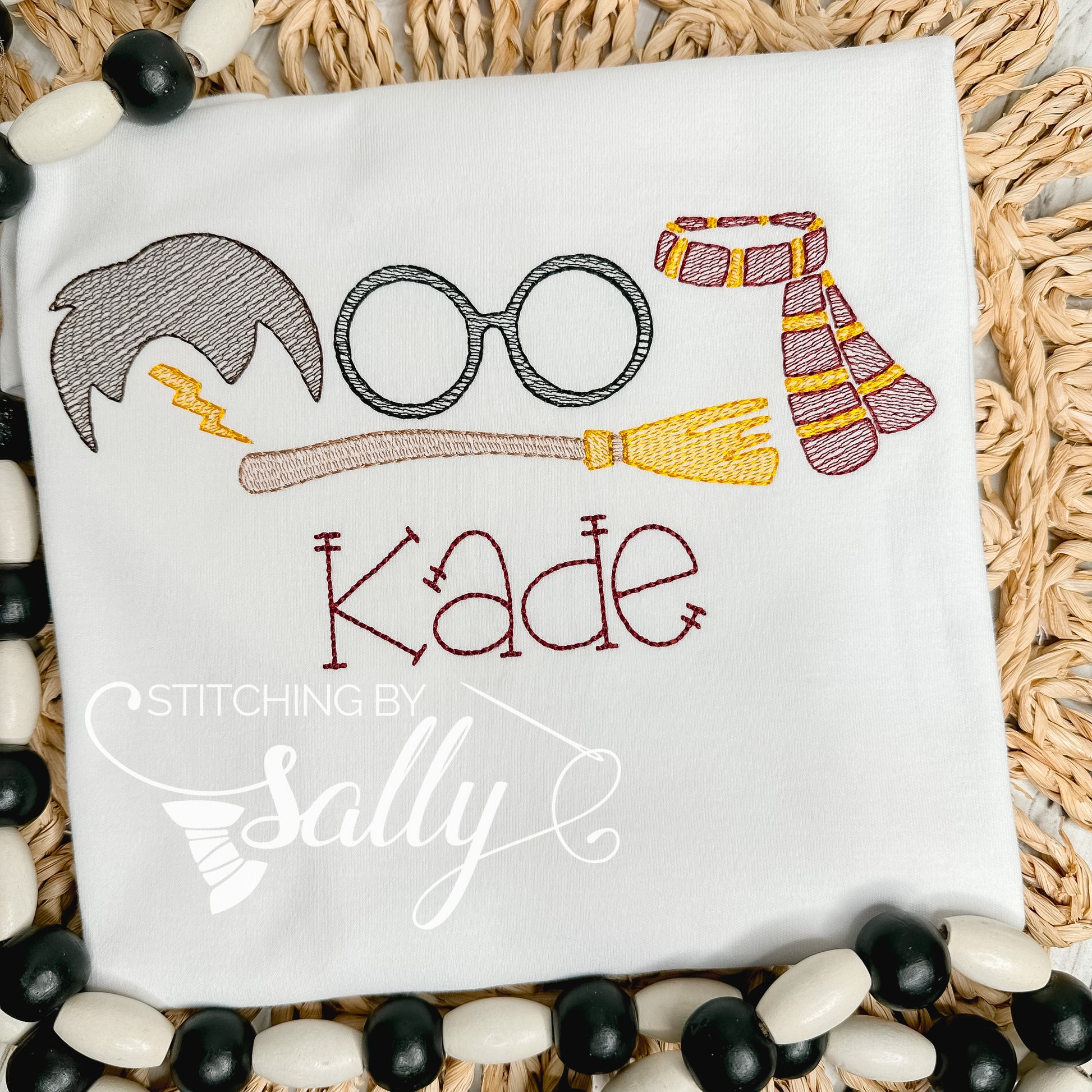 A white shirt with a few Harry Potter inspired designs embroidered on the shirt. A lighting bolt, glasses, scarf and a broom along with a name embroidered underneath. 
