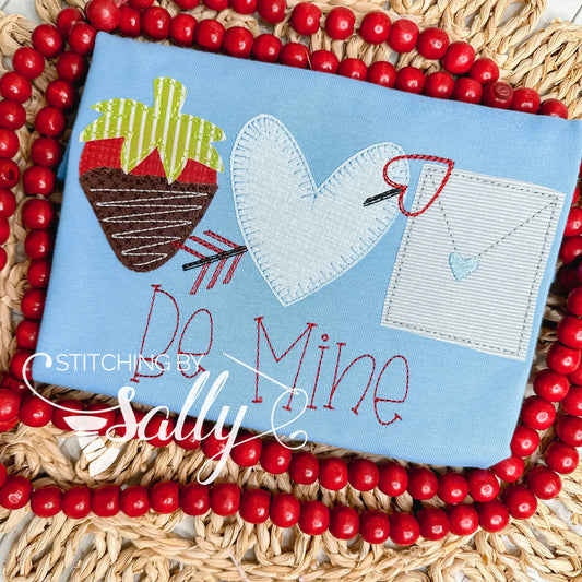 A strawberry, heart with arrow, and a letter are appliqué on a blue shirt. Be mine is embroidered underneath. 