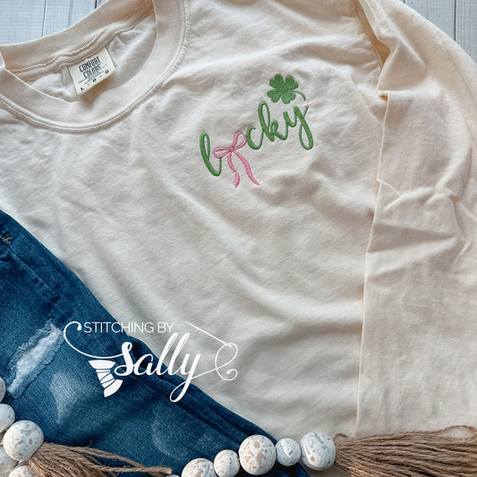 An ivory comfort colors shirt with the word lucky embroidered in vibrant green. The U is replaced with a pink bow for a girly look.