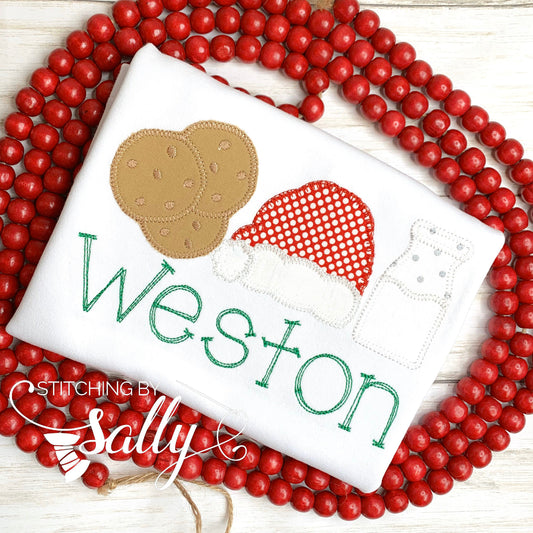 A white personalized shirt with green and red Christmas-themed embroidery, including a Santa hat and cookies, laying out on a surface with red beads.