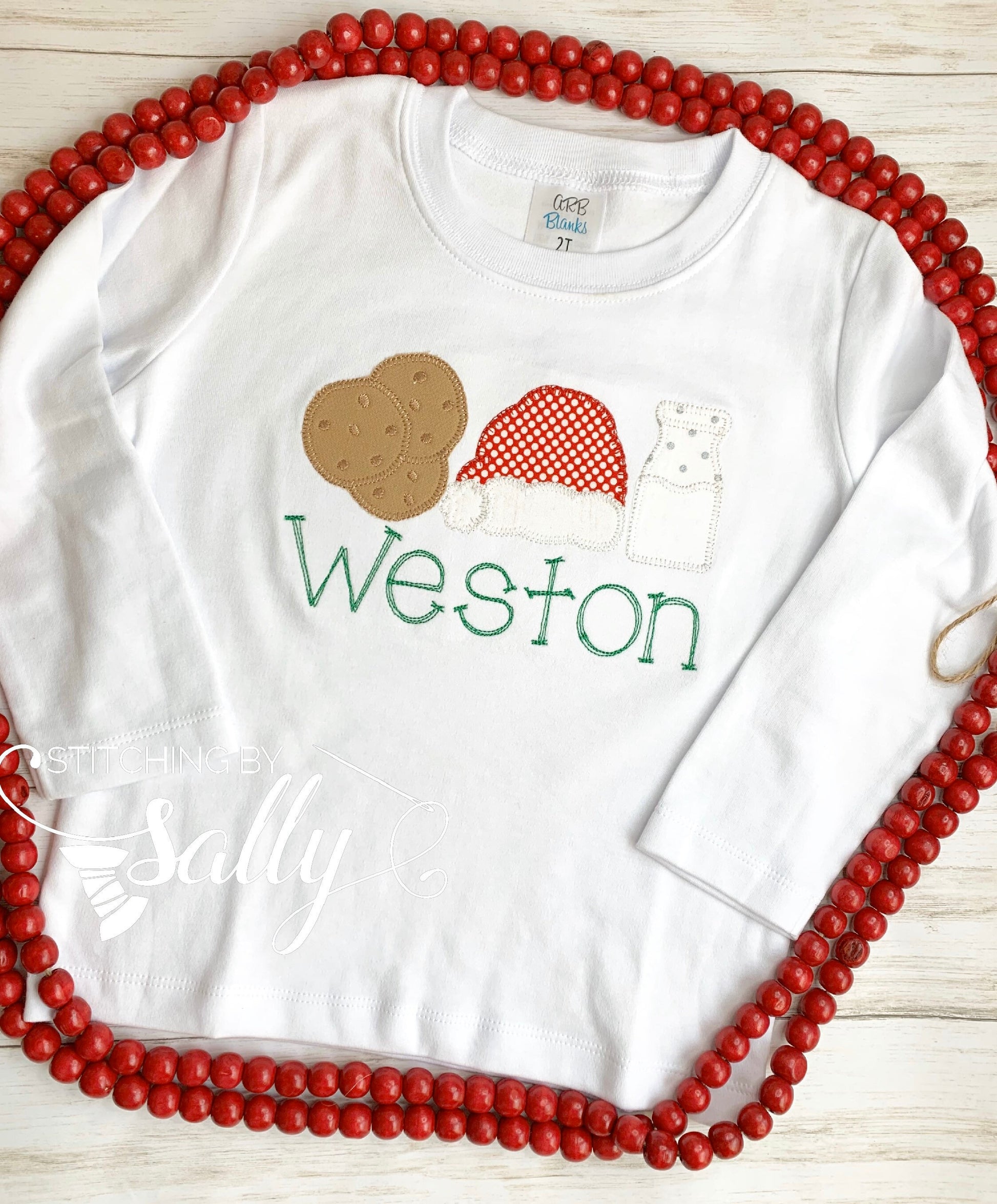 A white shirt laying flat with an applique of three christmas icons. Milk jug, cookies and a santa hat. 