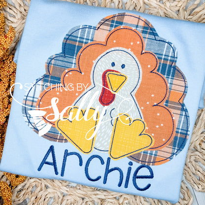 A boys' turkey applique shirt with a blue plaid background, featuring a yellow and white turkey design and the name 'Archie' sewn on the front.