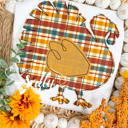 A toddler-sized shirt with a turkey applique design, featuring a plaid pattern and a personalized monogram, displayed on a bed of decorative rocks and flowers.