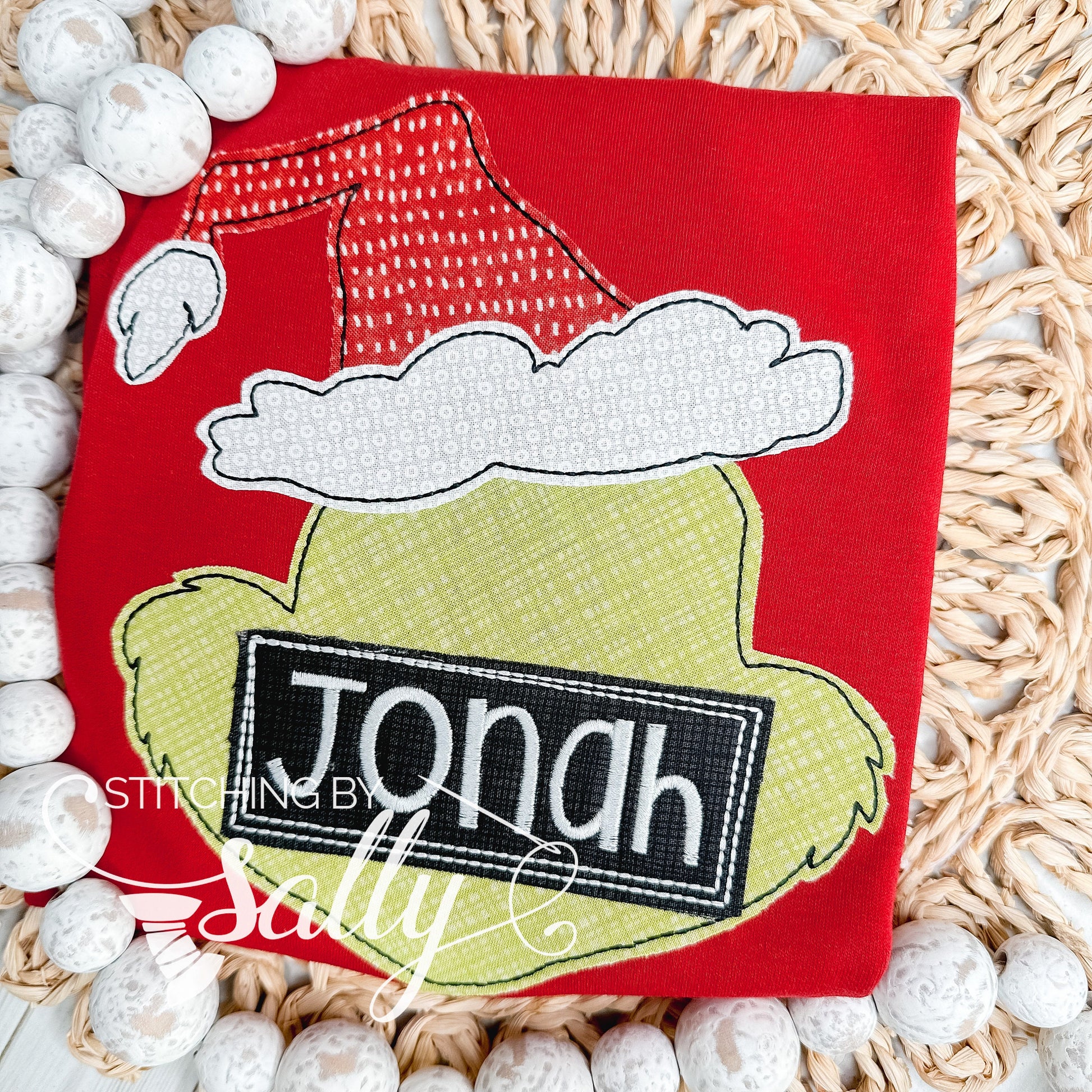 A red shirt with an inspired mean one design applique in green fabric with a red and white santa hat. Stitching by Sally
